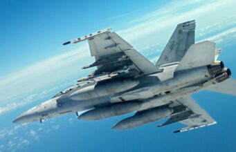 Two gray military fighter jets flying in close formation across a clear blue sky, with missiles mounted under the wings and contrails in the distance.