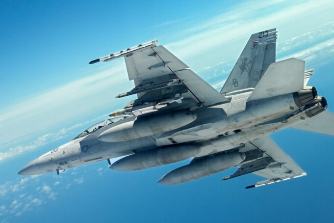 Boeing cleared to build long-range JDAM for US forces Two gray military fighter jets flying in close formation across a clear blue sky, with missiles mounted under the wings and contrails in the distance.