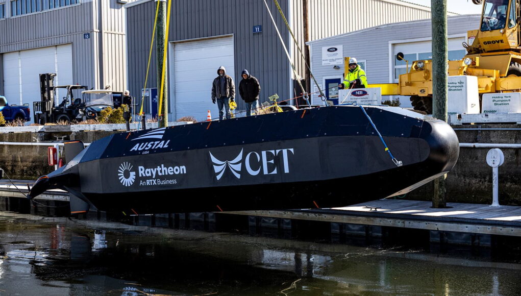 Lantern Fish Black submarine-shaped test vessel on a dock with workers nearby and construction equipment; logos Austal USA, Raytheon, and CET visible on the hull.