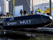 Black submarine-shaped test vessel on a dock with workers nearby and construction equipment; logos Austal USA, Raytheon, and CET visible on the hull.