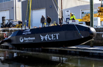 Raytheon tests drone submarine that launches weapons Black submarine-shaped test vessel on a dock with workers nearby and construction equipment; logos Austal USA, Raytheon, and CET visible on the hull.