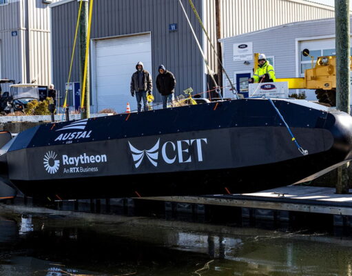 Black submarine-shaped test vessel on a dock with workers nearby and construction equipment; logos Austal USA, Raytheon, and CET visible on the hull.