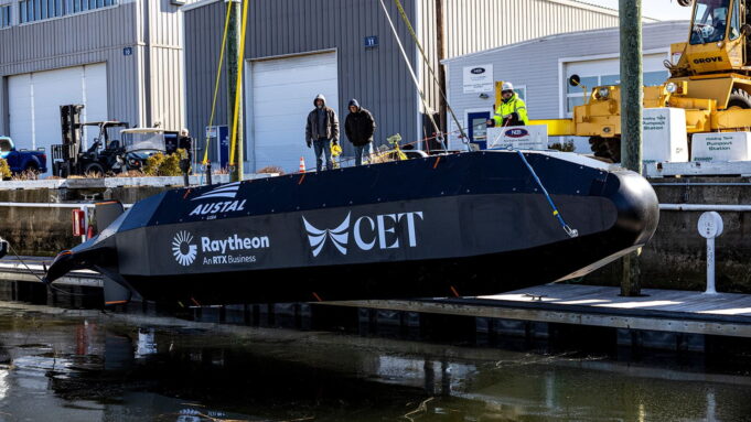 Raytheon tests drone submarine that launches weapons Black submarine-shaped test vessel on a dock with workers nearby and construction equipment; logos Austal USA, Raytheon, and CET visible on the hull.