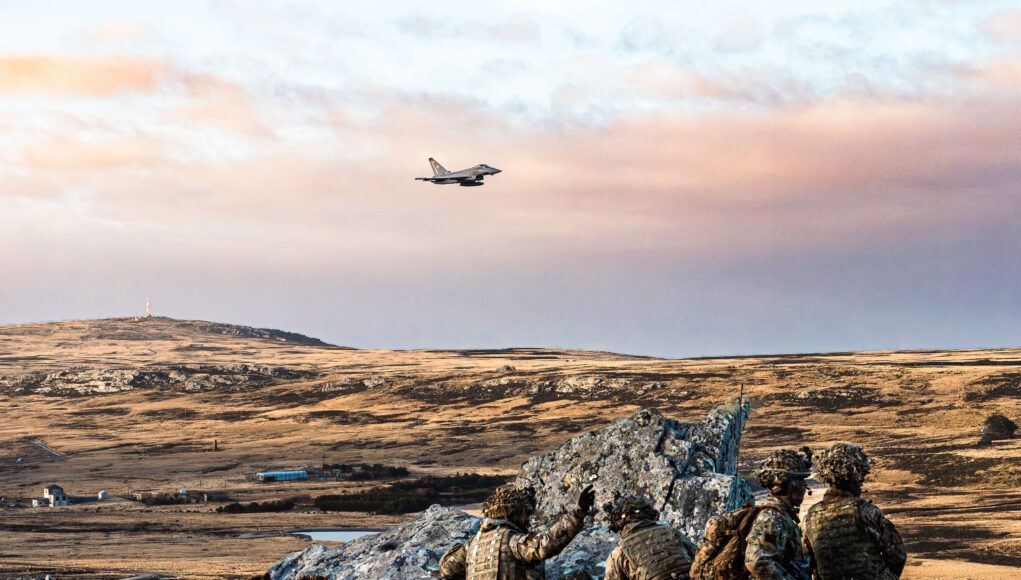 Exercise Cape Upholder Soldiers in camouflage crouch behind rocks on a barren landscape as a fighter jet flies overhead in the sky.
