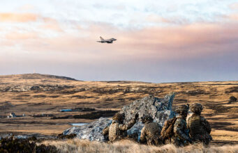 Troops and fighter jets in live fire exercise on Falklands Soldiers in camouflage crouch behind rocks on a barren landscape as a fighter jet flies overhead in the sky.