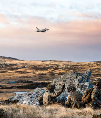 Soldiers in camouflage crouch behind rocks on a barren landscape as a fighter jet flies overhead in the sky.
