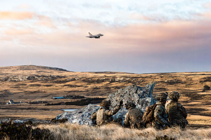 Soldiers in camouflage crouch behind rocks on a barren landscape as a fighter jet flies overhead in the sky.