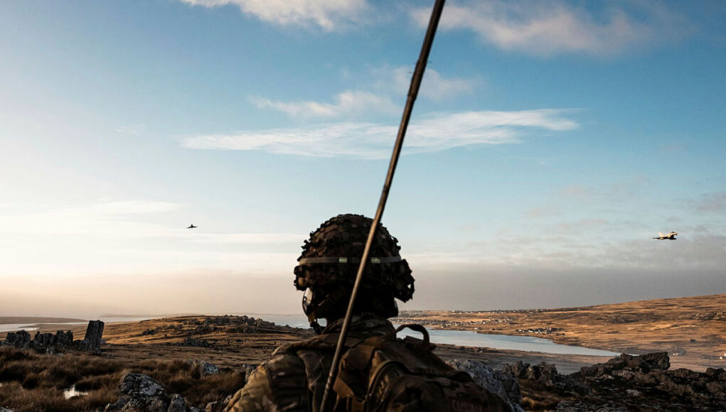 MPC_20260629_0191_0519.jpg.iCfptLSEMslPNwllYBMA.Vbhe4RBZPr Soldier in camouflage gear and helmet watches a barren, rocky landscape at sunset, with a helicopter flying in the distance.