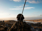 Soldier in camouflage gear and helmet watches a barren, rocky landscape at sunset, with a helicopter flying in the distance.