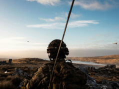 Army radio support deal to go to Babcock Soldier in camouflage gear and helmet watches a barren, rocky landscape at sunset, with a helicopter flying in the distance.