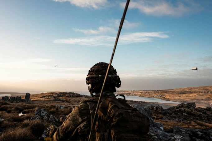 Soldier in camouflage gear and helmet watches a barren, rocky landscape at sunset, with a helicopter flying in the distance.