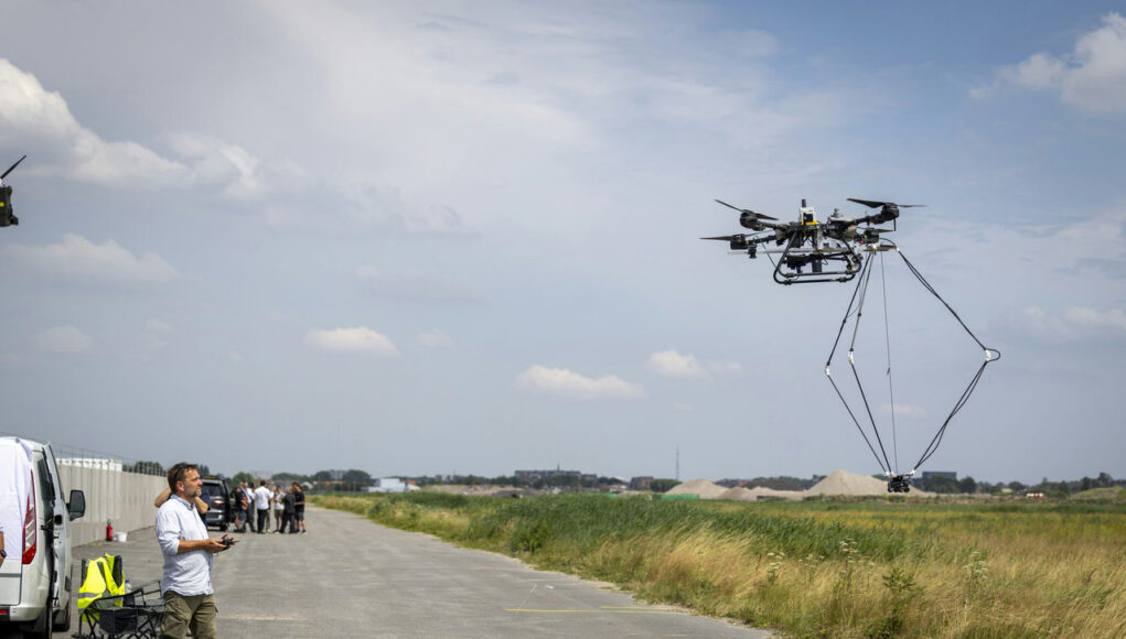 NATO Man operating a large multi-rotor drone hovering over a paved airfield beside a grassy field.