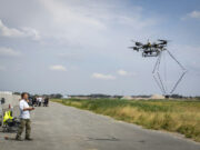 NATO tests autonomous drones without GPS Man operating a large multi-rotor drone hovering over a paved airfield beside a grassy field.