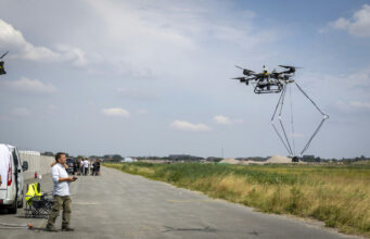 NATO tests autonomous drones without GPS Man operating a large multi-rotor drone hovering over a paved airfield beside a grassy field.