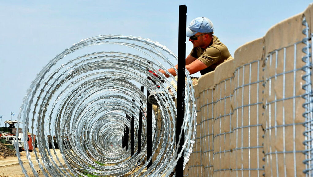 PJHQ-TRENTON-UNCLASS-20170904-021-003.jpg.iCfz5JaEMq_fBf1hYBMA.TvPIqNH4ch Construction worker in a cap and sunglasses installing coiled razor wire along a concrete border wall fence.