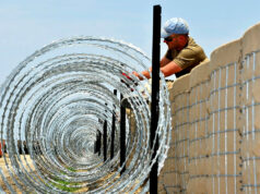 UK firms share $200m US barrier systems contract Construction worker in a cap and sunglasses installing coiled razor wire along a concrete border wall fence.