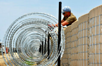 Construction worker in a cap and sunglasses installing coiled razor wire along a concrete border wall fence.