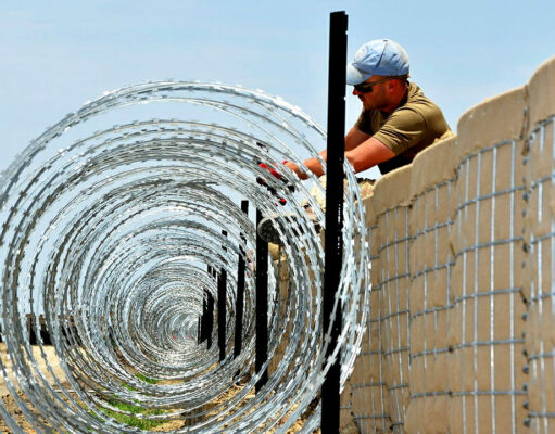 Construction worker in a cap and sunglasses installing coiled razor wire along a concrete border wall fence.
