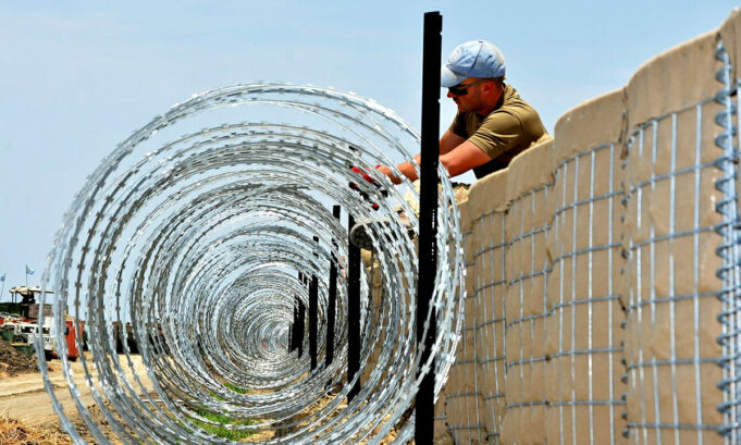 Construction worker in a cap and sunglasses installing coiled razor wire along a concrete border wall fence.