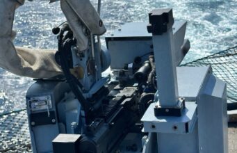 Close-up of a ship-mounted automatic weapon, with cables and sight, ocean in the background.