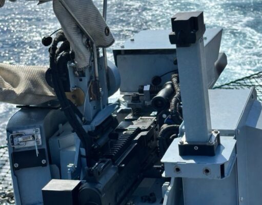Close-up of a ship-mounted automatic weapon, with cables and sight, ocean in the background.
