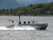 Gray cabin cruiser speeding across a bay with a tree-lined shoreline in the background, creating wake in the water.