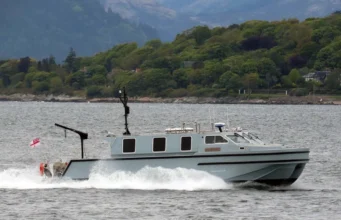 Gray cabin cruiser speeding across a bay with a tree-lined shoreline in the background, creating wake in the water.