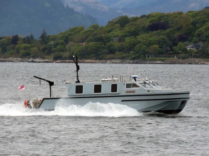 Navy survey team maps seabed in the Firth of Clyde Gray cabin cruiser speeding across a bay with a tree-lined shoreline in the background, creating wake in the water.