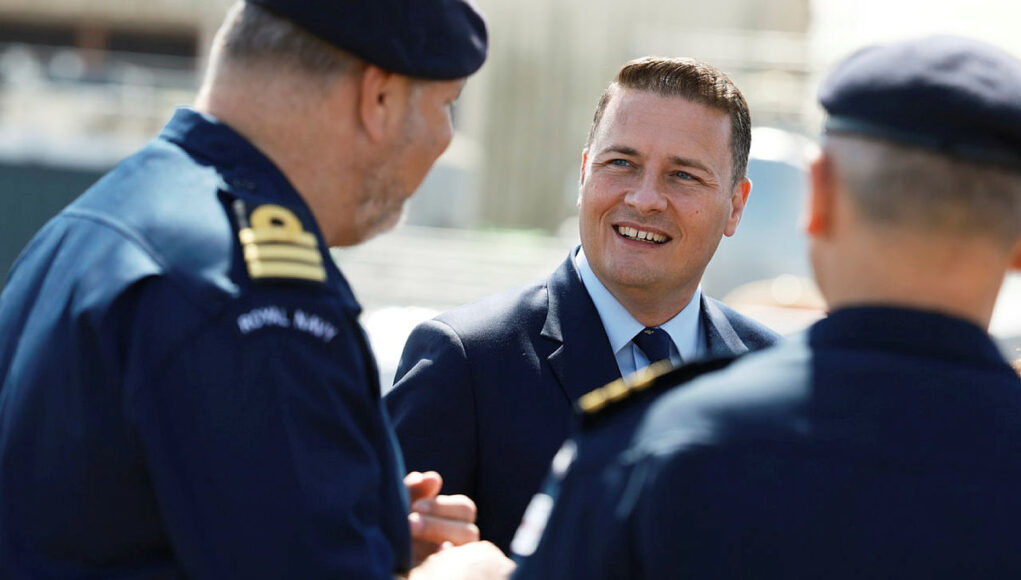 Man in a suit smiling and talking with two Royal Navy officers outdoors.