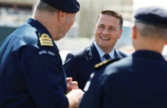 How Wes Streeting is faring as Defence Secretary Man in a suit smiling and talking with two Royal Navy officers outdoors.