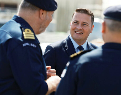 Man in a suit smiling and talking with two Royal Navy officers outdoors.