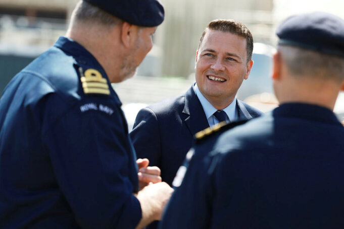 Man in a suit smiling and talking with two Royal Navy officers outdoors.