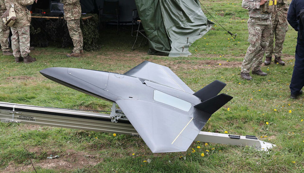 Sol26-060-146.jpg.iCfv9tOEMs2d4QVlYBMA.5uEe3gtCsh Unmanned military drone in a triangular, jet-like shape on a track grass field, with soldiers nearby and a tent in the background.