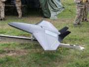 Britain brushes off threats from Russia Unmanned military drone in a triangular, jet-like shape on a track grass field, with soldiers nearby and a tent in the background.