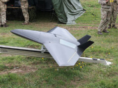 Britain brushes off threats from Russia Unmanned military drone in a triangular, jet-like shape on a track grass field, with soldiers nearby and a tent in the background.