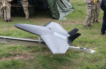 Unmanned military drone in a triangular, jet-like shape on a track grass field, with soldiers nearby and a tent in the background.