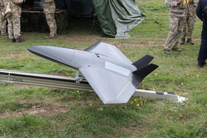 Unmanned military drone in a triangular, jet-like shape on a track grass field, with soldiers nearby and a tent in the background.