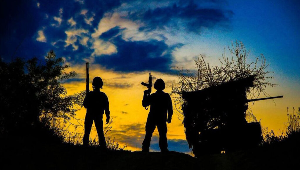 UKRAINE233 Two silhouetted figures with rifles stand on a hill at sunset, a ruined shack to the right against a blue‑orange sky.