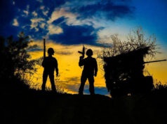 Russian claim to have taken Kostiantynivka disputed Two silhouetted figures with rifles stand on a hill at sunset, a ruined shack to the right against a blue‑orange sky.