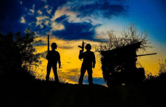 Russian claim to have taken Kostiantynivka disputed Two silhouetted figures with rifles stand on a hill at sunset, a ruined shack to the right against a blue‑orange sky.