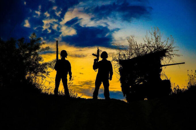 Russian claim to have taken Kostiantynivka disputed Two silhouetted figures with rifles stand on a hill at sunset, a ruined shack to the right against a blue‑orange sky.