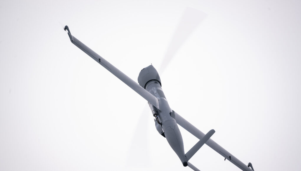 airbus-u030-flexrotor (1) View of a gray helicopter seen from below with spinning rotor blades against a pale sky.