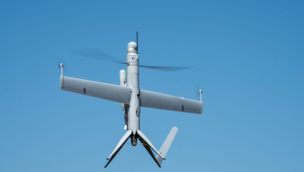Unmanned aerial vehicle (drone) with a fixed wing and helicopter-style rotor against a blue sky in flight.