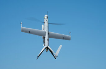 Unmanned aerial vehicle (drone) with a fixed wing and helicopter-style rotor against a blue sky in flight.