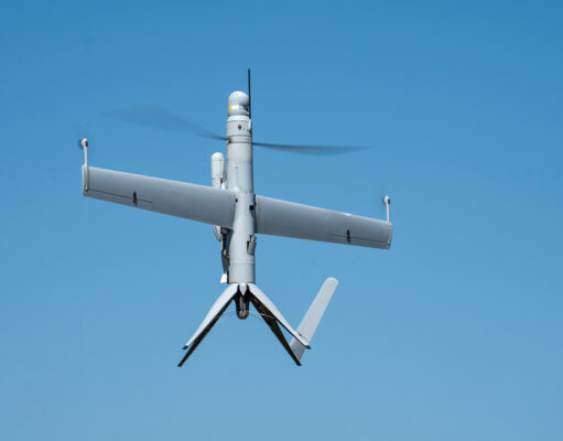 Unmanned aerial vehicle (drone) with a fixed wing and helicopter-style rotor against a blue sky in flight.