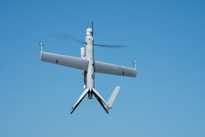 Unmanned aerial vehicle (drone) with a fixed wing and helicopter-style rotor against a blue sky in flight.