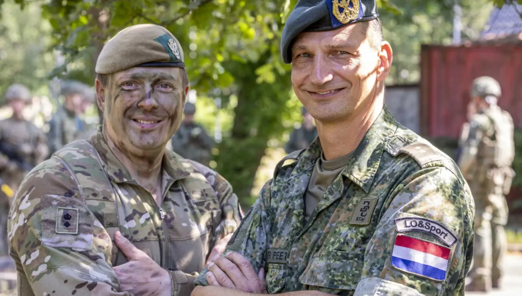 army007-official-20260715-070-0186 Two soldiers in camouflage shake hands and smile, with more troops visible in the background outdoors.