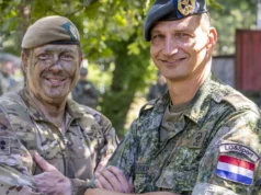 Two soldiers in camouflage shake hands and smile, with more troops visible in the background outdoors.