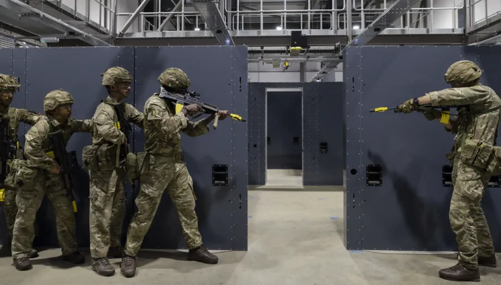 Five soldiers in camouflage gear practice room-clearing behind gray modular walls in an indoor training facility.