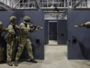 Five soldiers in camouflage gear practice room-clearing behind gray modular walls in an indoor training facility.